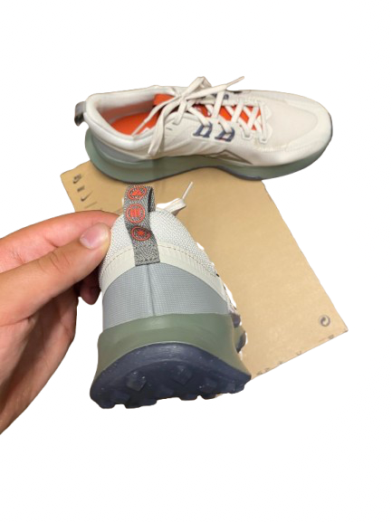 A pair of white trail running shoes with gray and olive green accents, shown with one shoe held by a hand to display the sole and heel, and the other placed on a cardboard backing against a white background.
