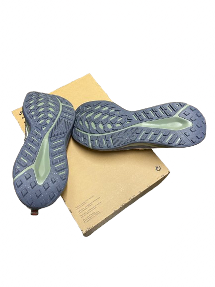 A pair of gray trail running shoes with a light green midsole accent, shown from the sole view resting on a tan cardboard box against a white background.
