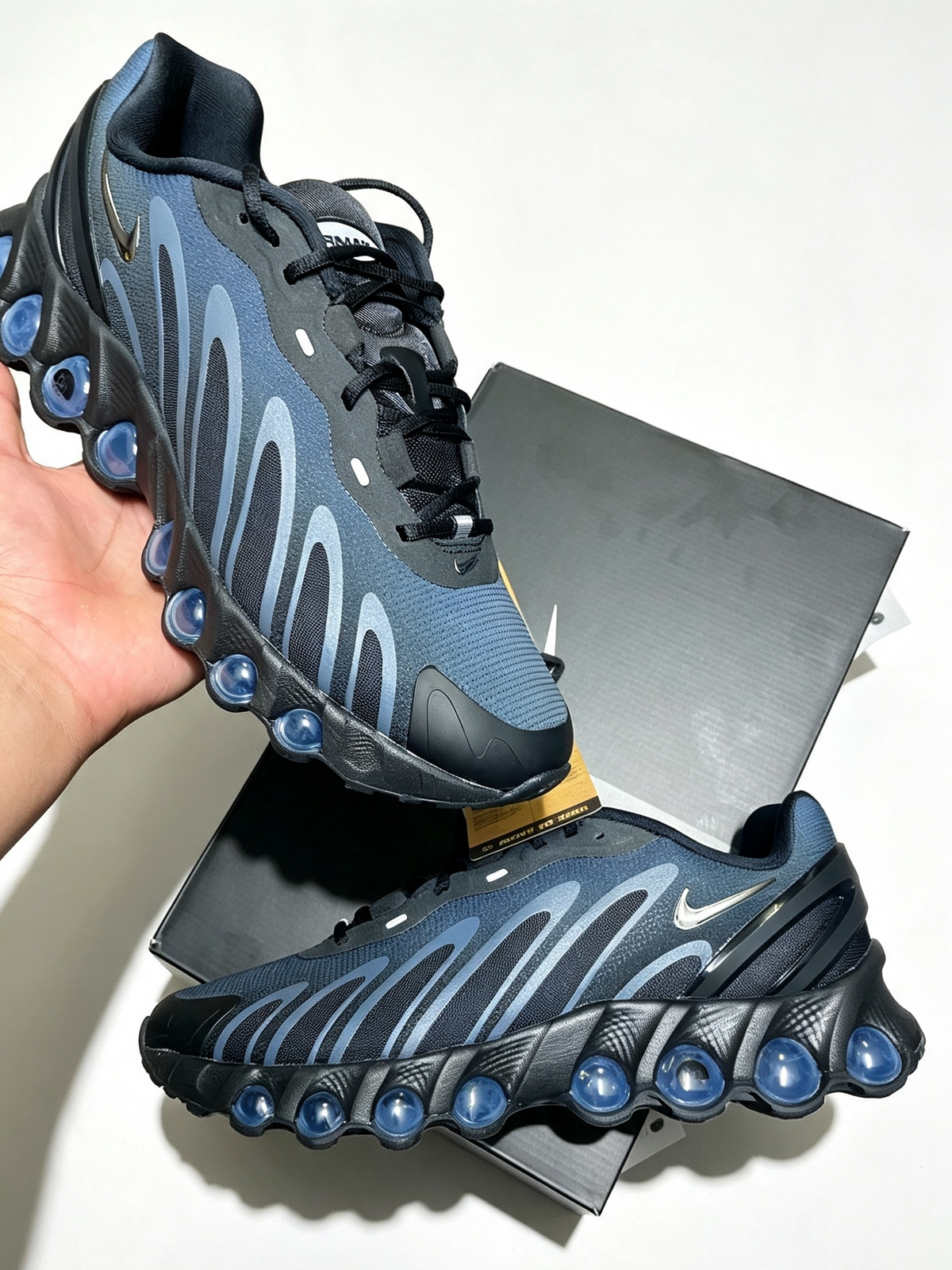 A pair of Nike running shoes in navy blue and black with light blue wave-like accents, held by a hand with the shoebox in the background.
