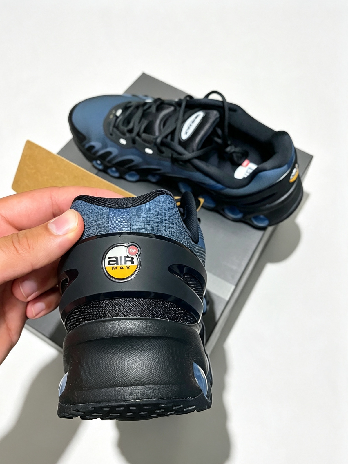 A close-up of a hand holding the rear half of a black and navy Air Max sneaker, with the full shoe visible in the background on a gray box against a white wall.
