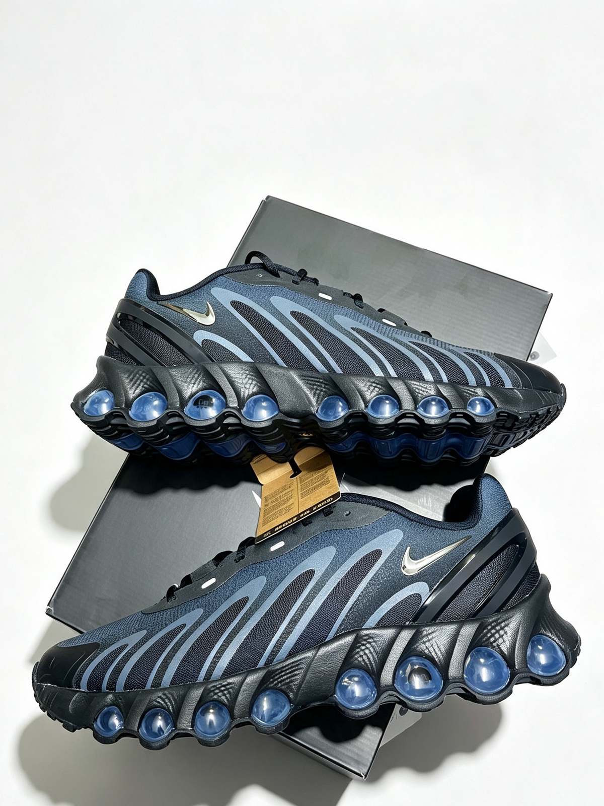 A pair of Nike Air Max dn8 sneakers in dark navy blue and black with metallic silver wave accents and blue translucent air pods, displayed on a white background with their gray shoebox.
