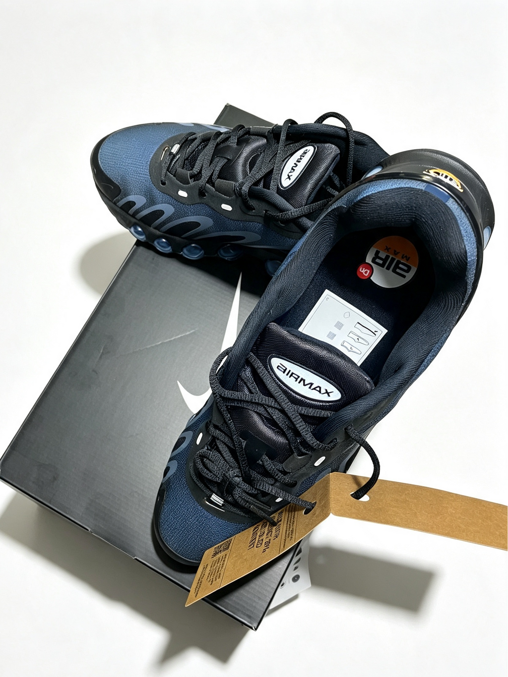A pair of Nike Air Max 80s sneakers in navy blue and black, displayed on top of their dark gray shoebox with a paper hangtag. The shoes feature the signature Air Max logo on the tongue and a yellow Nike+ logo on the collar.