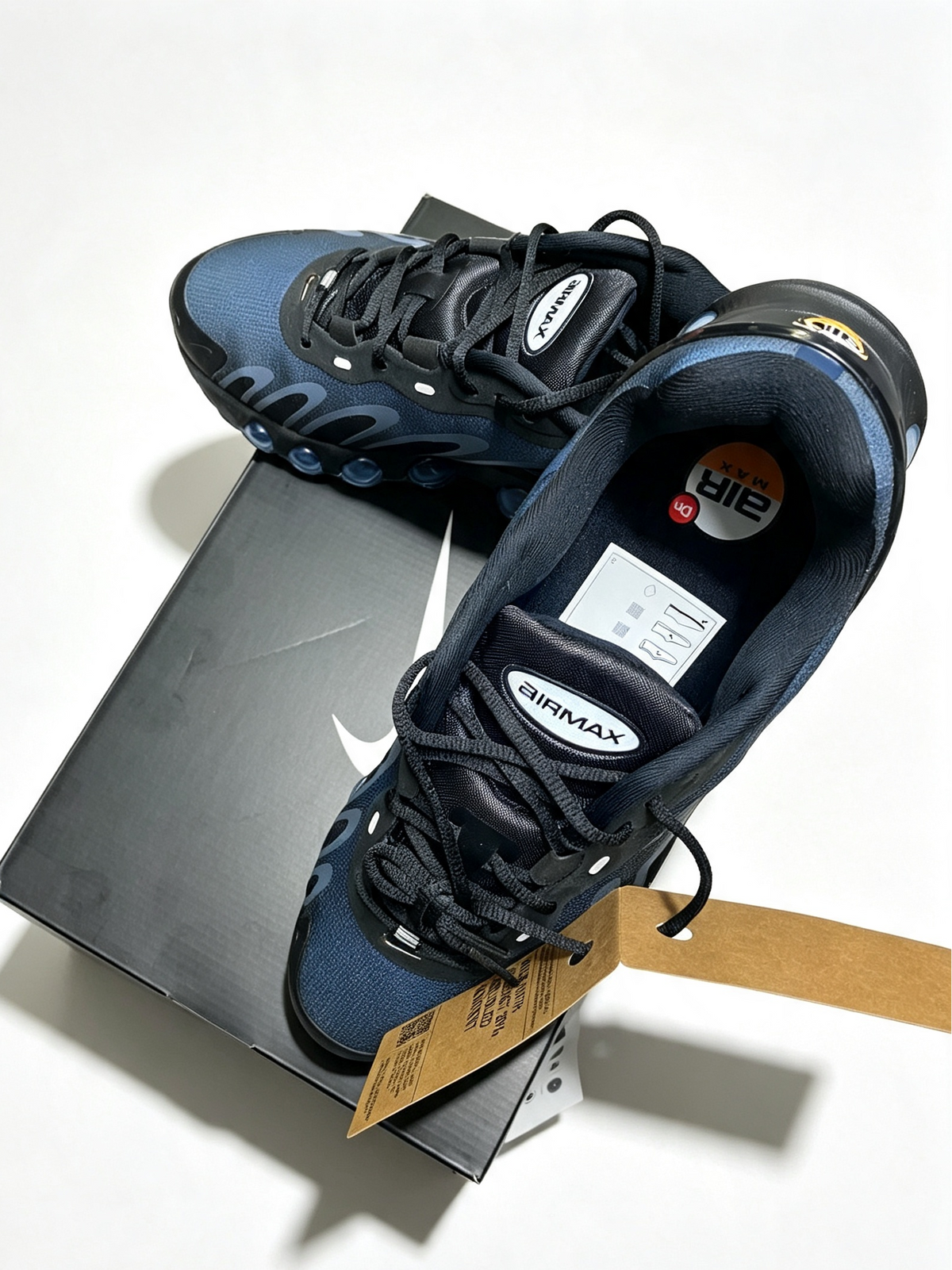 A pair of Nike Air Max 80s sneakers in navy blue and black, displayed on top of their dark gray shoebox with a paper hangtag. The shoes feature the signature Air Max logo on the tongue and a yellow Nike+ logo on the collar.